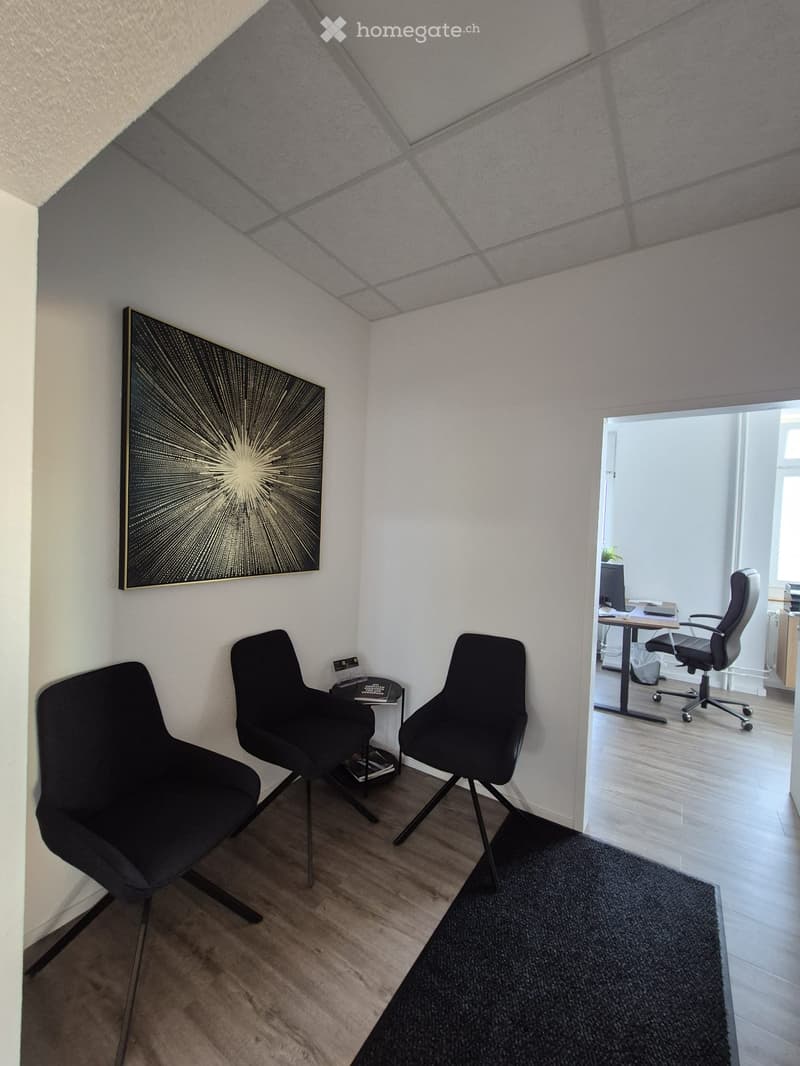 A modern, minimalist room with three black chairs, a small table, and a large abstract artwork on the wall. The room has light wooden flooring and a black rug.