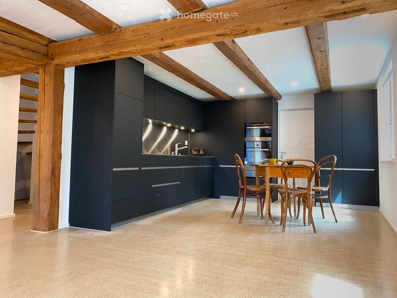 Modern kitchen with dark cabinetry, stainless steel appliances, and exposed wooden beams. Includes a small dining area with a wooden table and chairs.