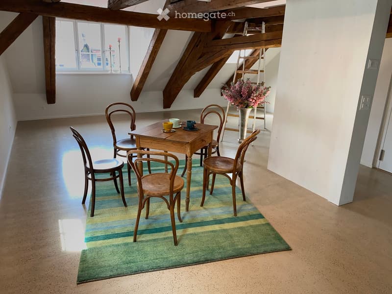 Bright attic room with wooden beams, a small wooden table surrounded by chairs, a vase with flowers, and a colorful striped rug.