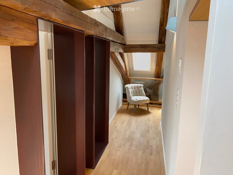 A narrow hallway with wooden flooring, white walls, and a single cream-colored upholstered chair placed near a small window at the end.