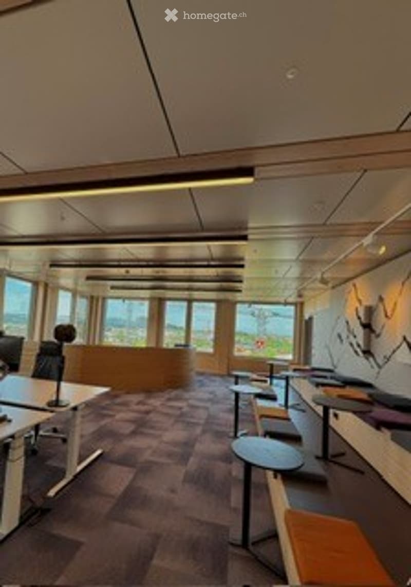 Modern office space with large windows, wooden desks, chairs, and stools arranged in rows.