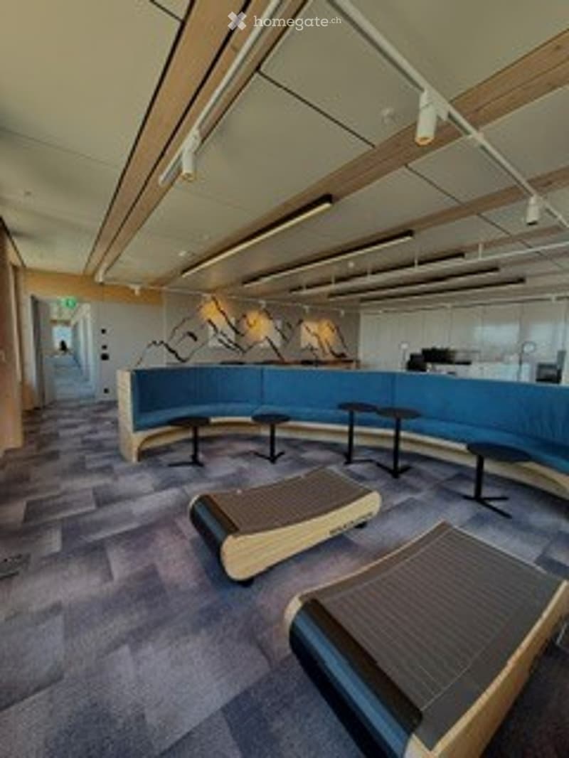 Modern lounge area with blue curved sofa, small tables, and wooden chairs. The space features contemporary design with a mix of wood and blue upholstery.