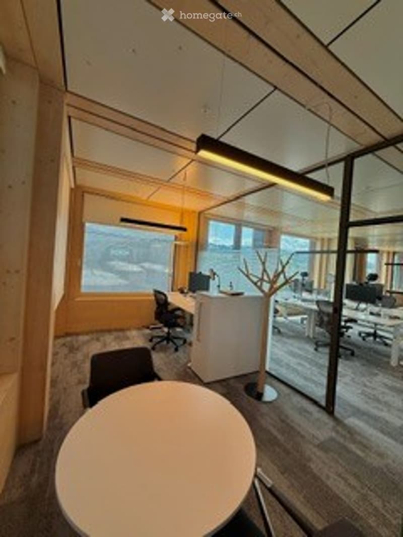 Modern office space with large windows offering a scenic view, featuring desks, chairs, a round table, and a screen.