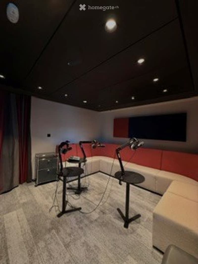 Modern, soundproofed room with grey carpet, white walls, and red accent wall. Features a drum set, microphone stands, a sofa, a small table, and a chair.