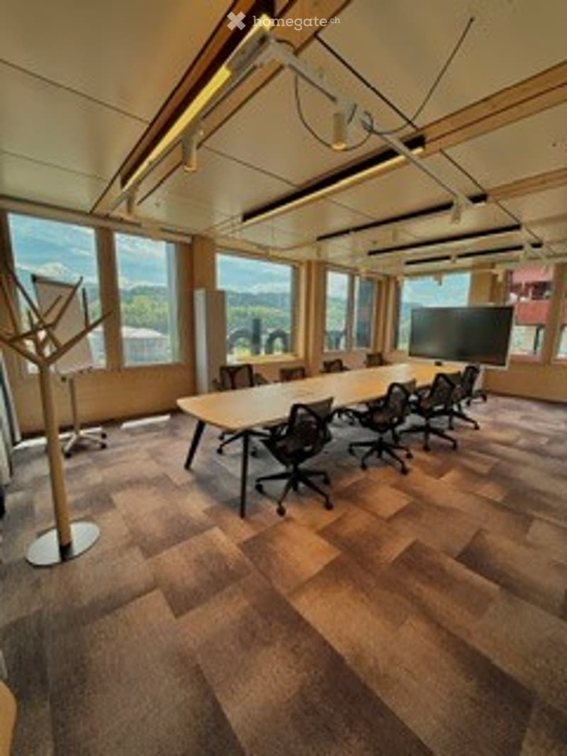 Modern conference room with large windows offering a scenic view, a long conference table surrounded by office chairs, a large screen at one end, and a coat rack near the entrance.