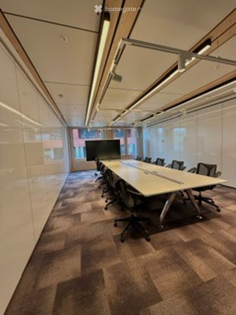 Modern conference room with a long rectangular table, several office chairs, and large windows allowing natural light. The room features a carpeted floor and recessed lighting.