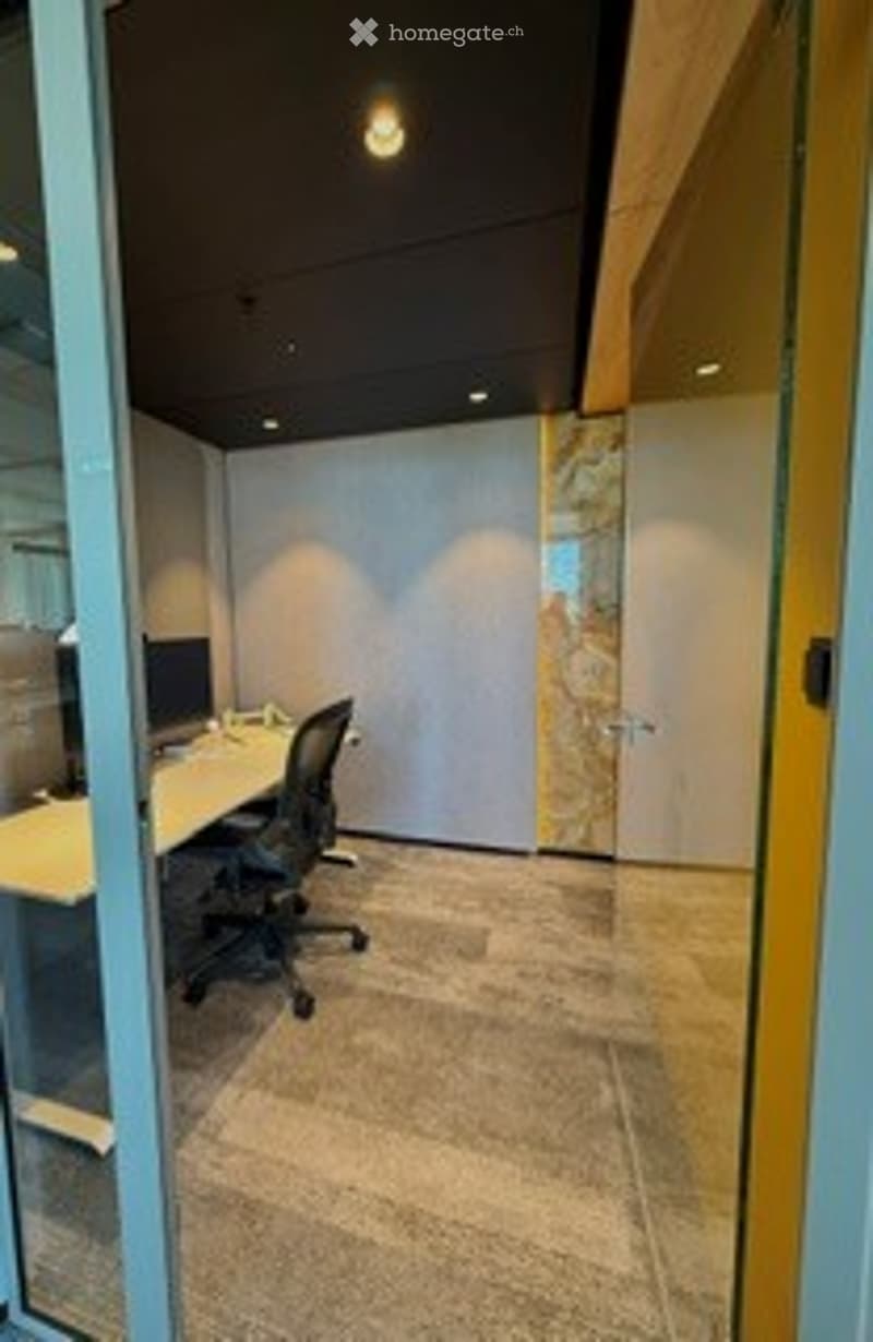 Modern office space with a sleek white desk, ergonomic chair, and computer monitor. The room features contemporary decor with a large glass wall, recessed lighting, and a neutral color palette.
