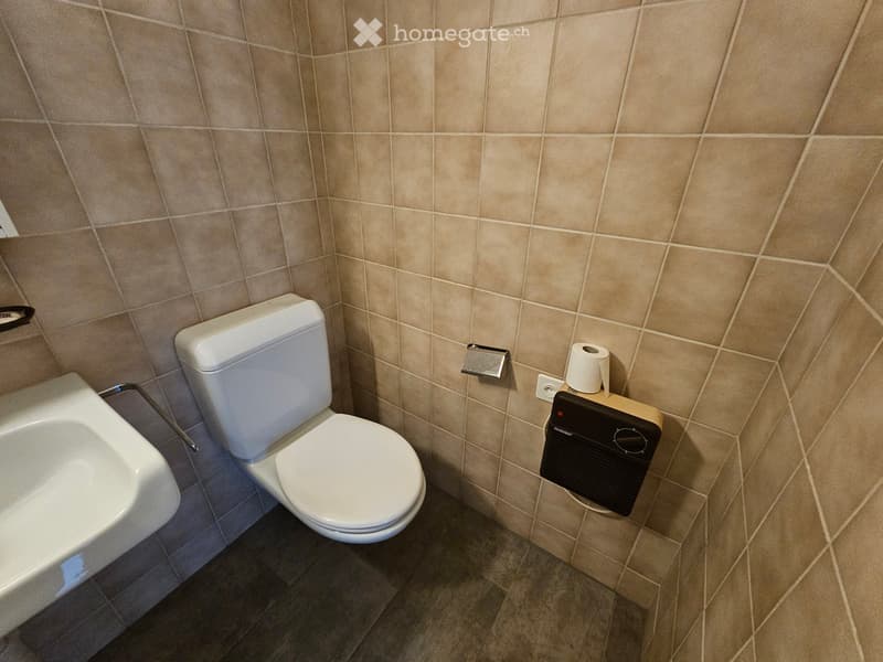 Modern bathroom with beige tiled walls, a white toilet, a sink, and a towel holder with toilet paper. The floor is tiled in a darker shade.