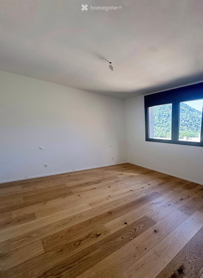 Empty room with light-colored wooden flooring and white walls, featuring a large window with a view of trees and mountains.