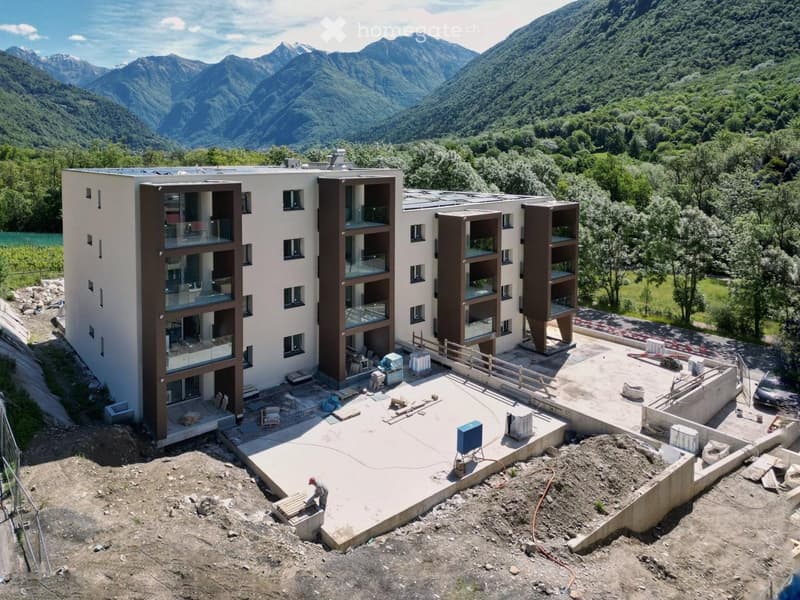 Modern multi-story building with large glass balconies, surrounded by a construction site with materials and machinery, set against a backdrop of mountains and greenery.