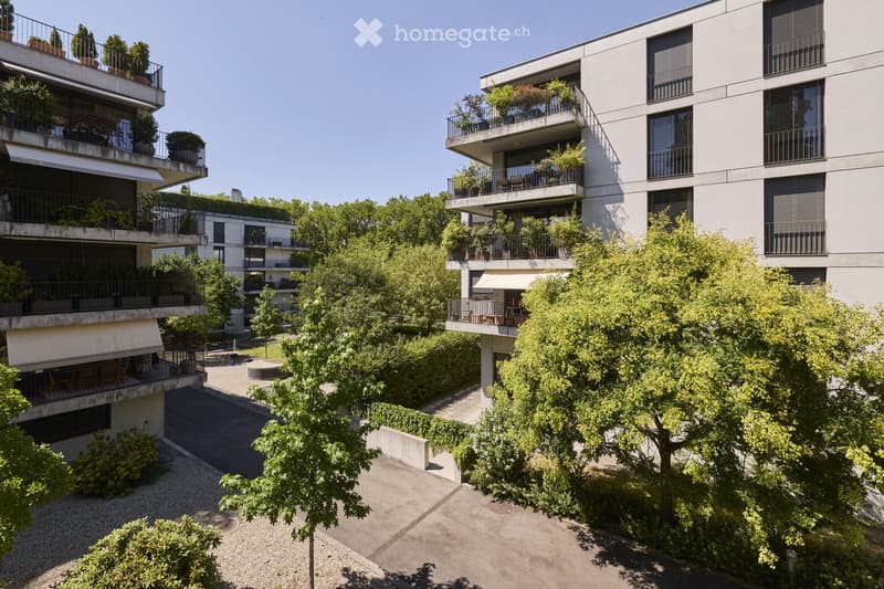 Modern multi-story residential building with balconies, greenery, and trees in the courtyard.