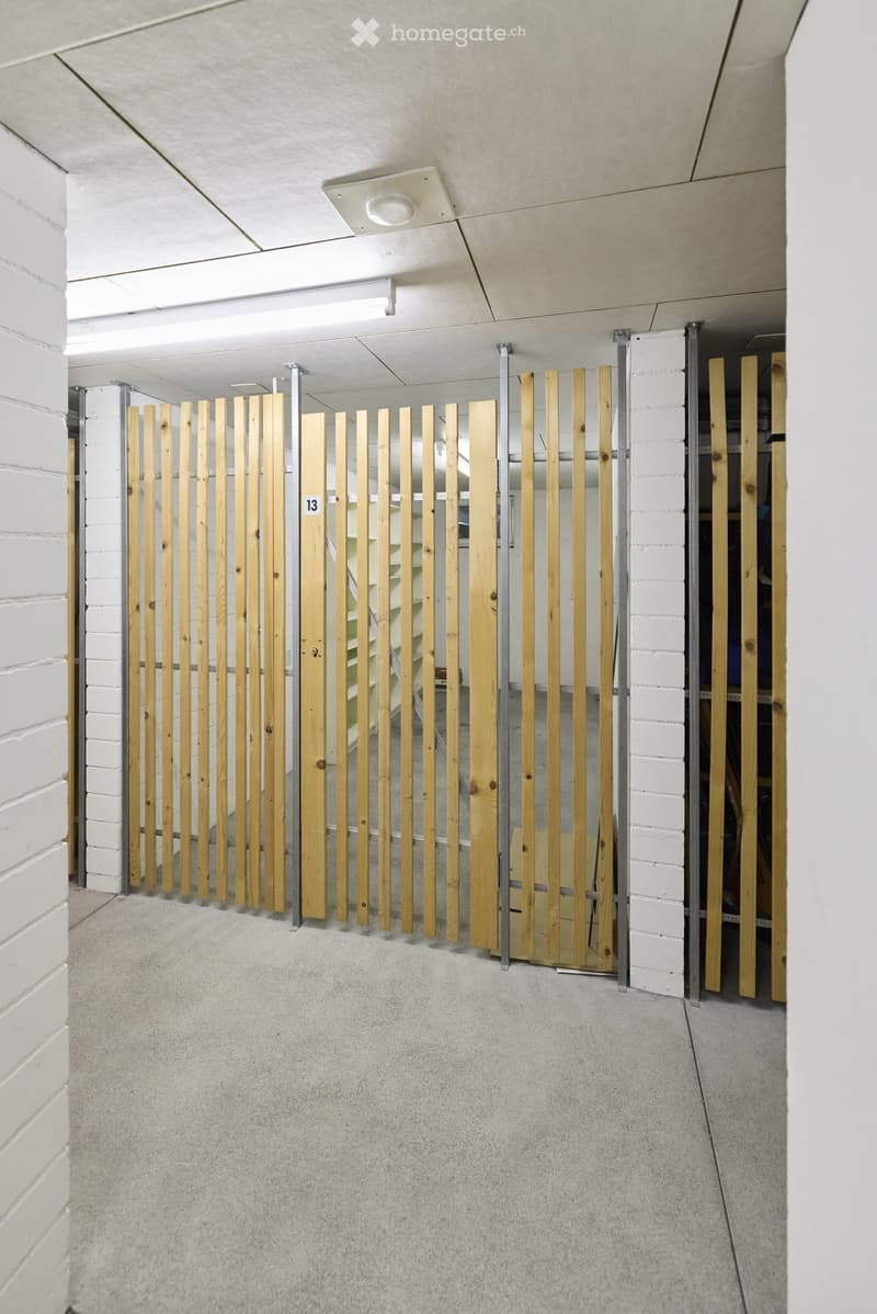 A modern hallway with white brick walls, a concrete floor, and a wooden slatted gate with the number 13 displayed on it.