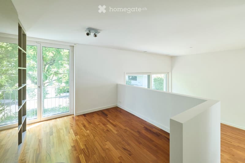 An empty room with wooden flooring, white walls, large windows, and a sliding glass door leading to a balcony.