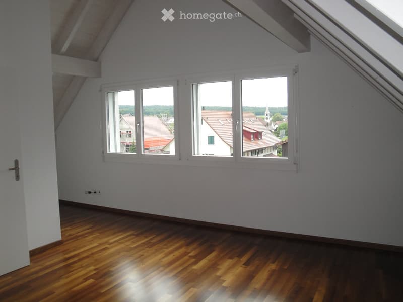Empty attic room with wooden flooring, white walls, and large windows offering a view of the neighborhood.