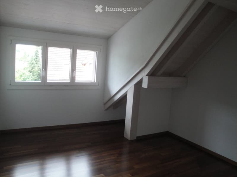 Empty hallway with wooden flooring, white walls, and a staircase leading upwards. Three windows on one side allow natural light to enter.
