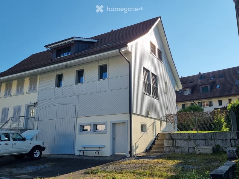 Two-story house with white siding, brown roof, multiple windows, garage, and small garden area.