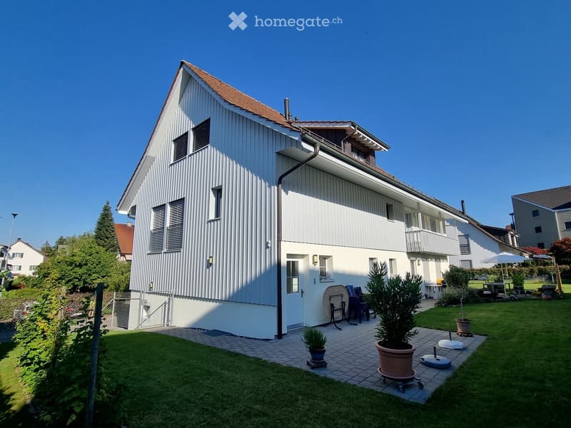 Two-story house with white siding, multiple windows, and a small balcony on the upper level. The front yard features a well-maintained lawn, potted plants, and outdoor furniture.