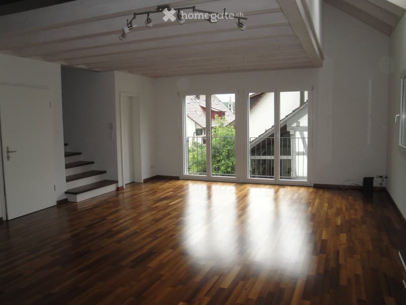 Empty room with wooden flooring, large windows, and a staircase leading upwards.