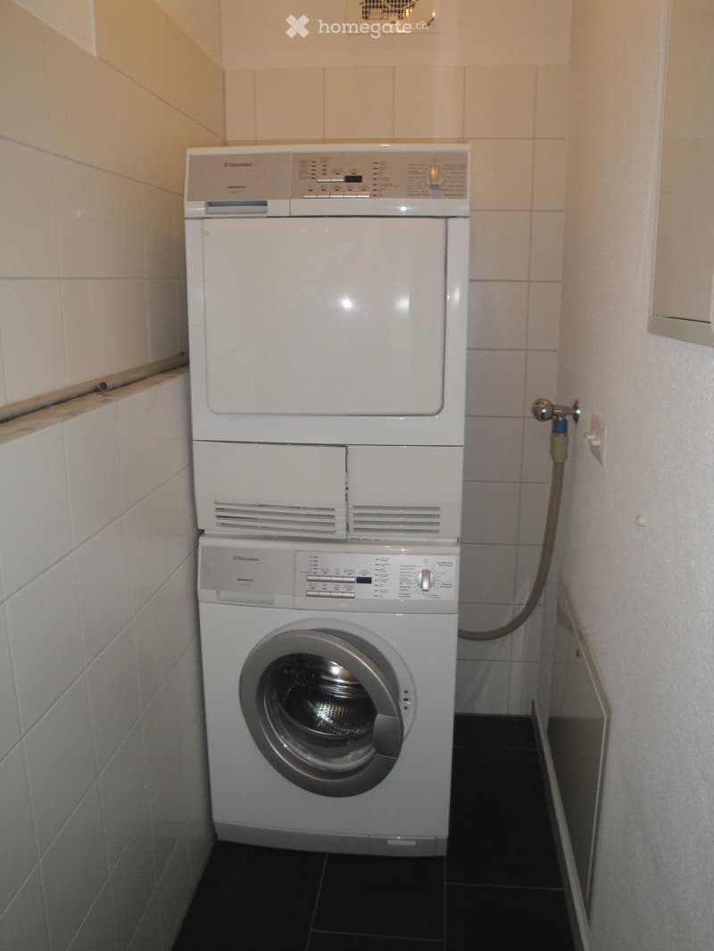 A compact bathroom with a stacked washing machine and dryer unit, white tiled walls, and a dark tiled floor.