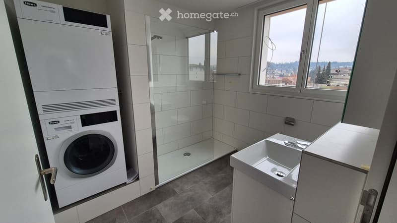 Modern bathroom with a glass-enclosed shower, a white sink, and a washing machine. The space features tiled walls and a large window offering a view of the surroundings.
