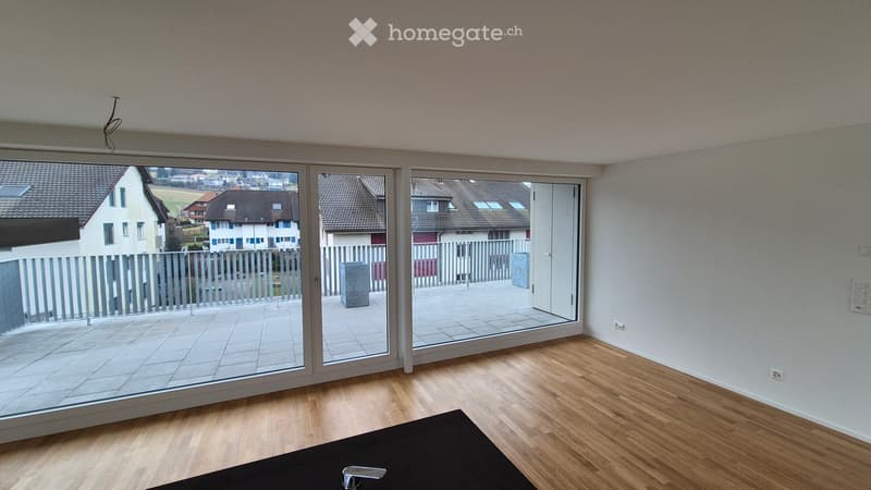 Empty room with wooden flooring, large sliding glass doors leading to a balcony, and a view of the neighborhood.