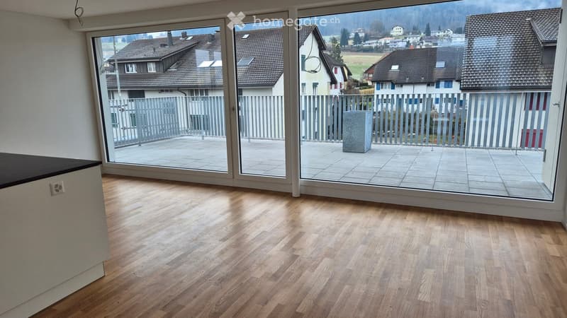 Empty room with wooden flooring, large glass sliding doors leading to a balcony with a view of houses and a fenced area.
