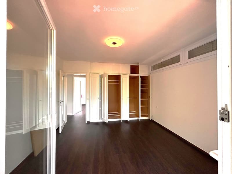 Empty hallway with wooden flooring, white walls, and built-in white closet units with open doors revealing empty shelves. A ceiling light fixture is visible.