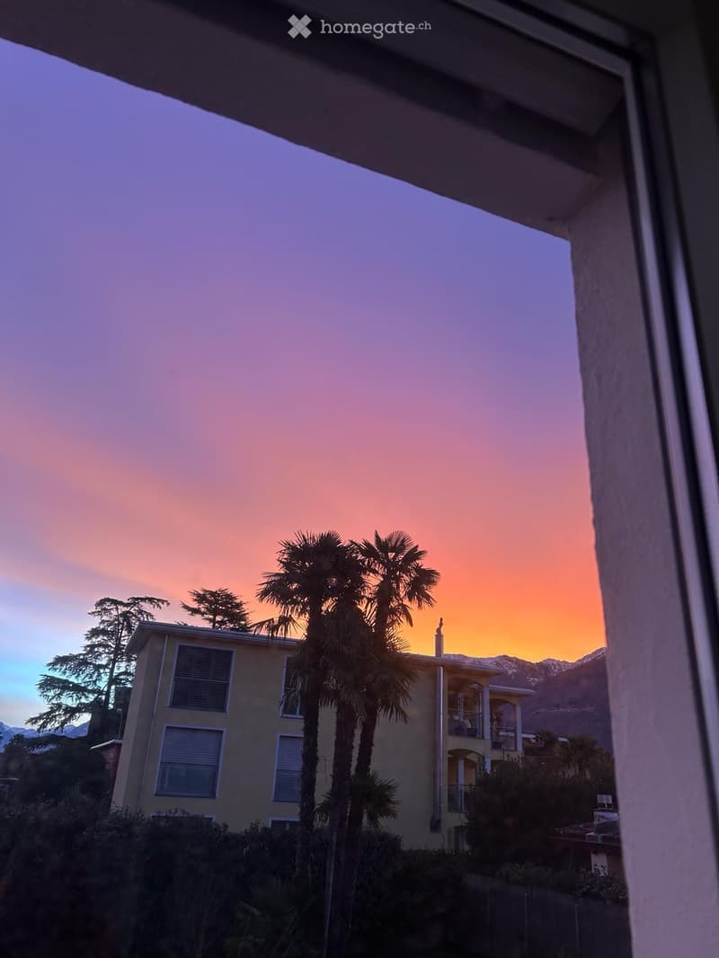 A two-story house with a balcony, surrounded by palm trees and mountains, viewed during sunset.
