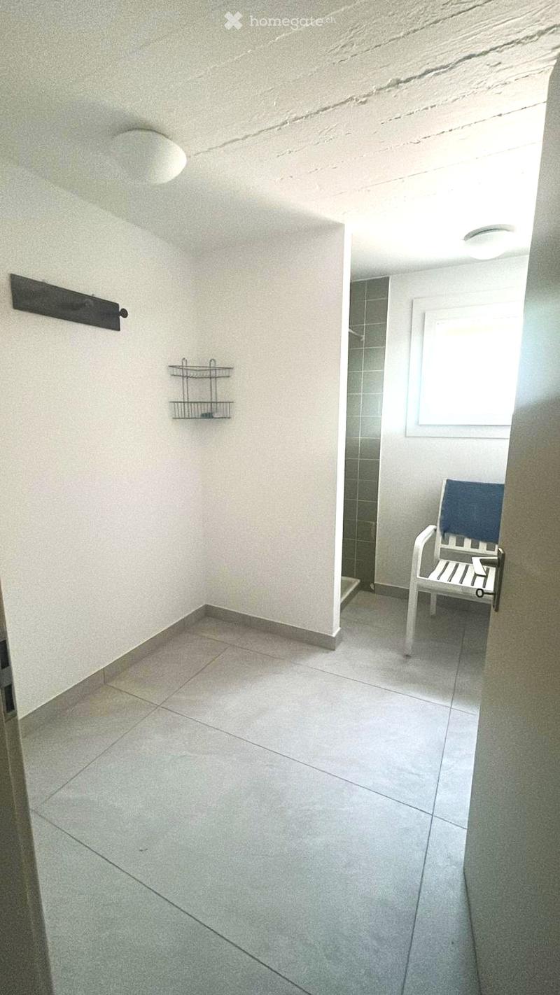 Balcony with tiled floor, white walls, a small shower area, a white plastic chair with a blue cushion, and two wall-mounted shelves.