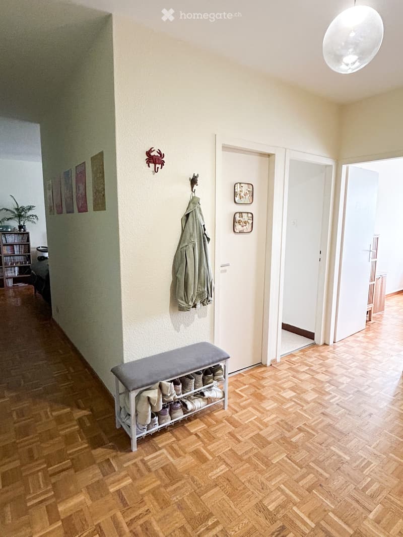 Hallway with wooden flooring, a shoe rack with multiple pairs of shoes, a coat hanging on the wall, and a small bench with a cushioned seat.