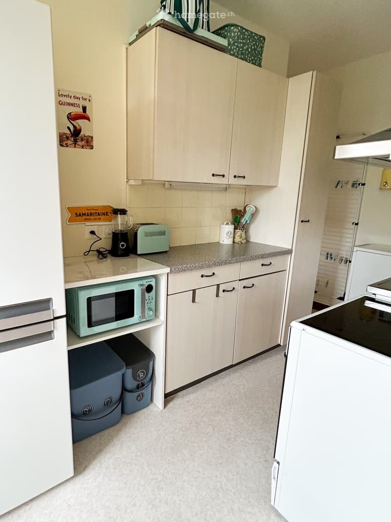 A compact kitchen with white cabinets, a light green microwave, a toaster, a blender, and a white oven. The countertop has a few small items, and there are two blue trash bins under the microwave.