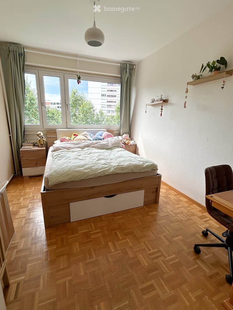 A well-lit bedroom with wooden flooring, a large window with curtains, a bed with pillows and blankets, a chair, a small table, and a shelf with decorative items.