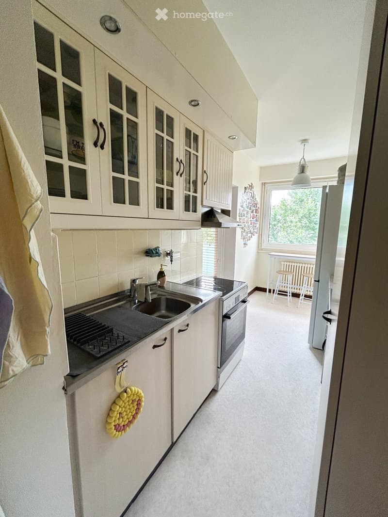 Bright kitchen with white cabinets, stainless steel sink, oven, and a small dining area with stools and a table. A window offers a view of greenery outside.