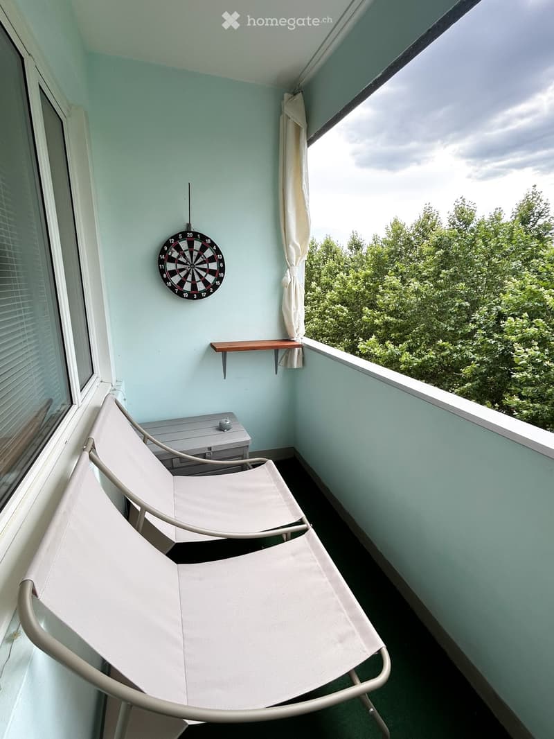 Balcony with two white lounge chairs, a small table, and a dartboard mounted on the wall, overlooking a green, tree-filled area.
