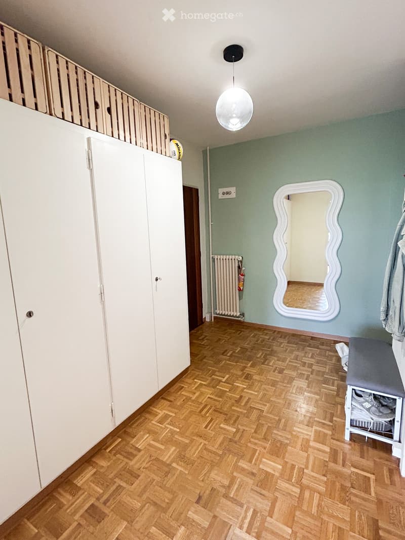 Empty room with wooden herringbone flooring, white built-in closet, a round mirror with a white frame, a radiator, and a small bench with shoes on it.