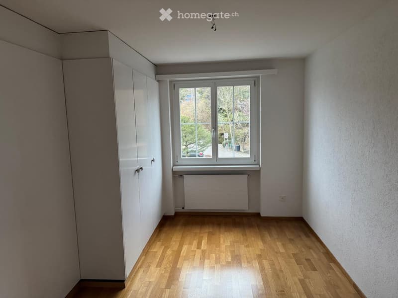 Empty room with wooden flooring, white walls, built-in closet, window with radiator below, and visible outdoor view.