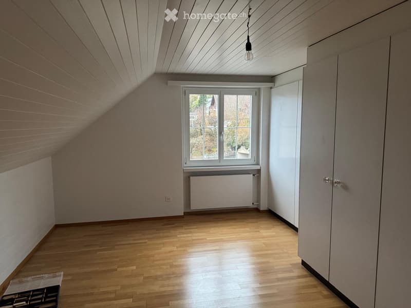 Empty attic room with wooden floors, slanted ceilings, white walls, and a window with a radiator below it.