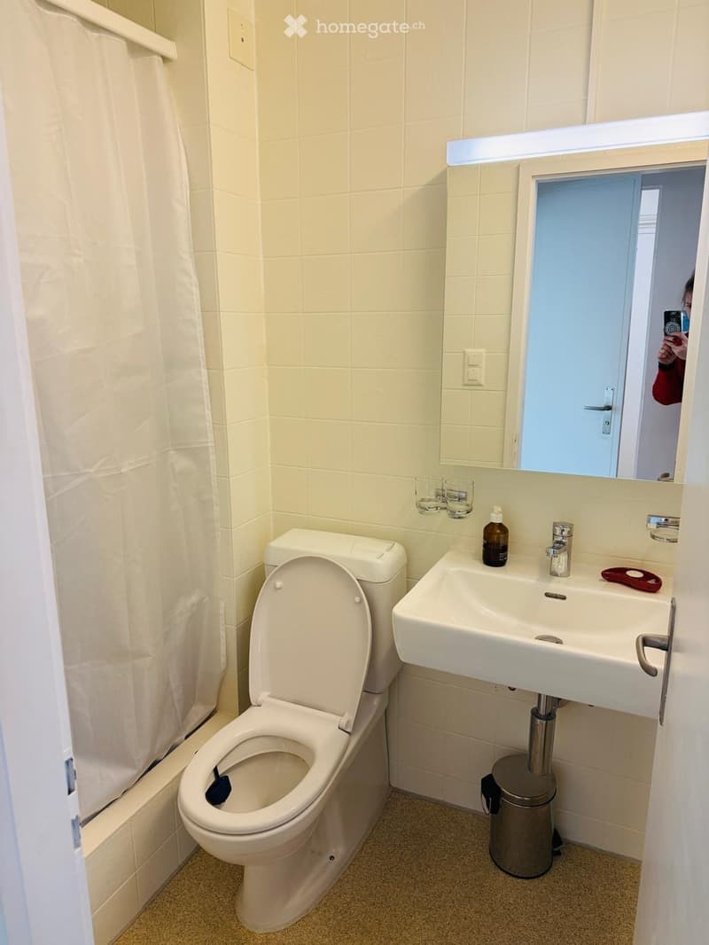 Compact bathroom with white tiled walls, a toilet, a sink with a mirror above it, and a small trash can. The sink has a bottle and a cup on it.