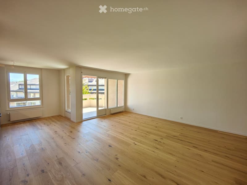 Empty room with light wooden flooring, white walls, large windows, and sliding glass doors leading to a balcony.