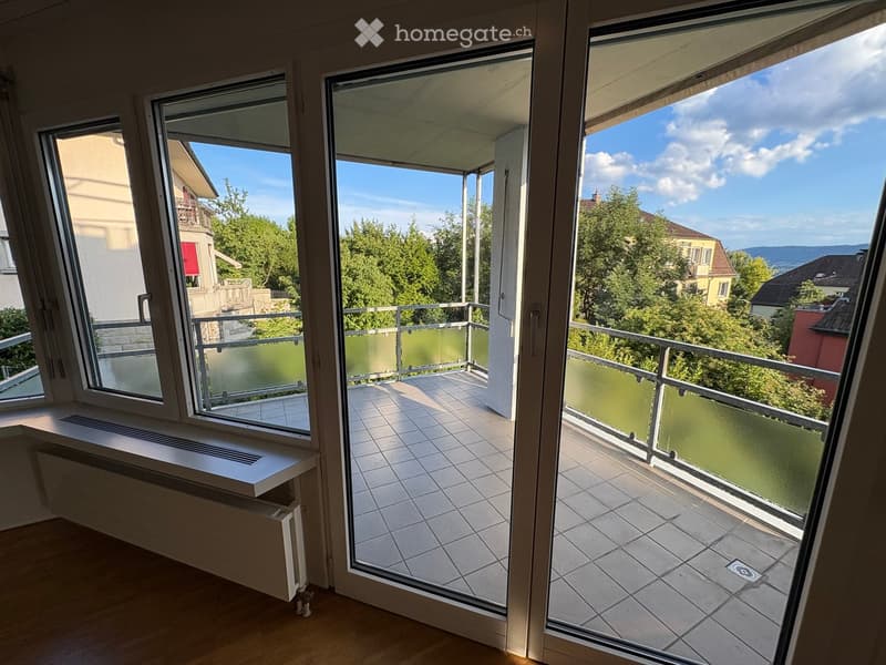 Balcony with tiled floor, glass railing, and scenic view of trees and buildings.