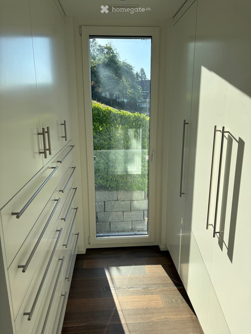A modern hallway with light-colored walls, wooden flooring, and built-in white cabinets with metal handles. A large glass door at the end leads to a garden view.