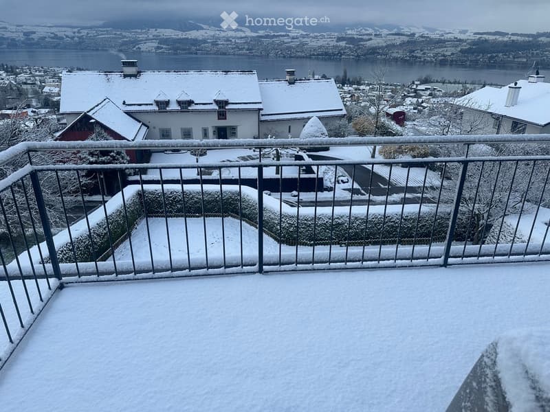 Snow-covered balcony overlooking a town with houses, a lake, and distant mountains.