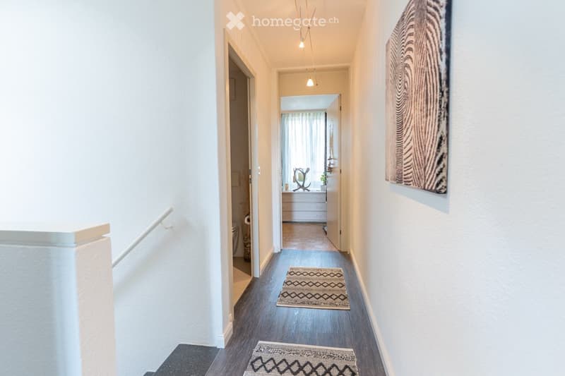 The image shows a hallway with a hardwood floor, white walls, and recessed lighting fixtures. There is a patterned rug on the floor and a decorative wall hanging visible. The hallway leads to a room with a window visible at the end.