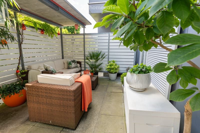 Covered patio with wicker furniture, potted plants, and a tiled floor. The patio is surrounded by a slatted fence and has a lush, green plant overhanging the space.