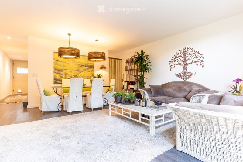 Spacious open-plan living and dining area with hardwood floors, white walls, large windows, two pendant light fixtures, a sofa, coffee table, and a dining table with chairs. The room also features a fireplace and a wall decor with a tree motif.