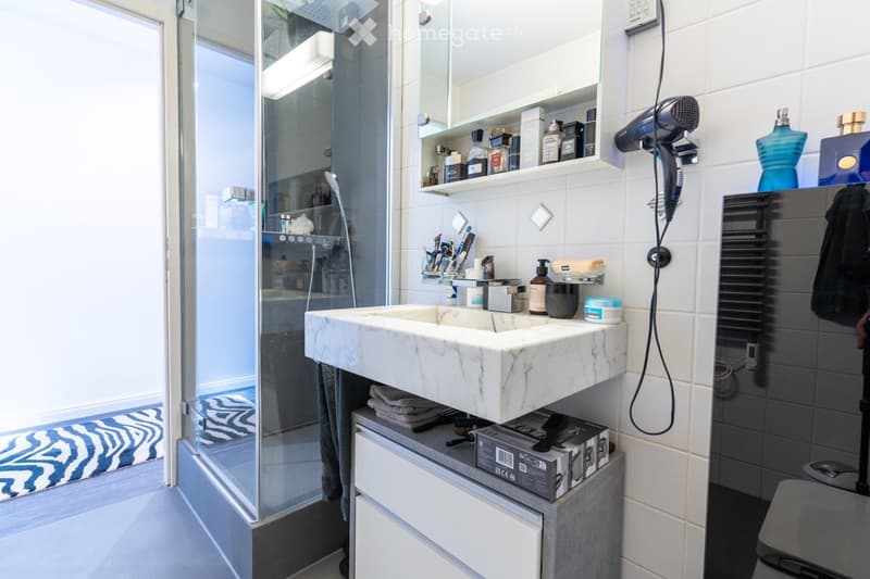 The image shows a modern bathroom with a marble vanity, shelving above the vanity, and a glass shower enclosure. The bathroom appears to be well-equipped with various personal care items and accessories visible on the shelves and vanity.