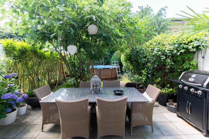Lush, green garden with a variety of plants and trees, an outdoor dining area with a stone table and wicker chairs, a Buddha statue, and a small above-ground swimming pool in the background.