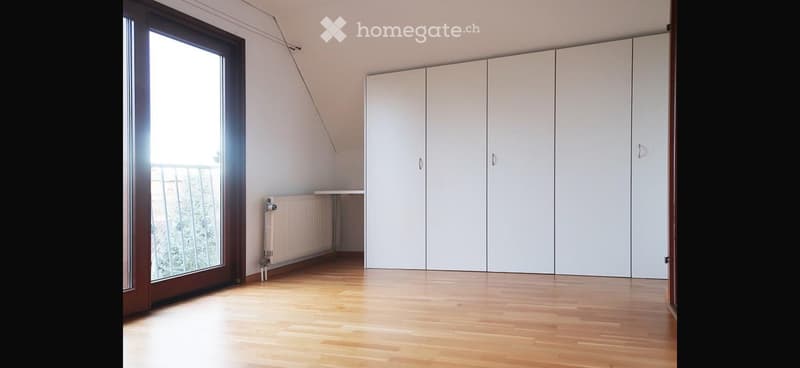 Empty room with wooden flooring, white built-in wardrobes, large window with balcony access, radiator, and a small table.