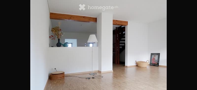 Empty room with wooden flooring, white walls, and a staircase leading upwards. There is a floor lamp, a small shelf with a vase and a watering can, and a woven basket on the floor.