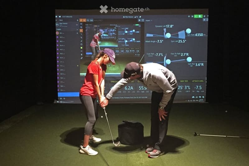 Two individuals practice golf on an indoor simulator, using a golf club and observing data on a large screen.